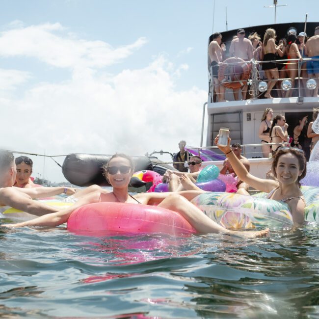 People are enjoying a sunny day on the water, lounging on colorful inflatable floats. A boat in the background has more people gathered on it. The atmosphere is festive and relaxed.