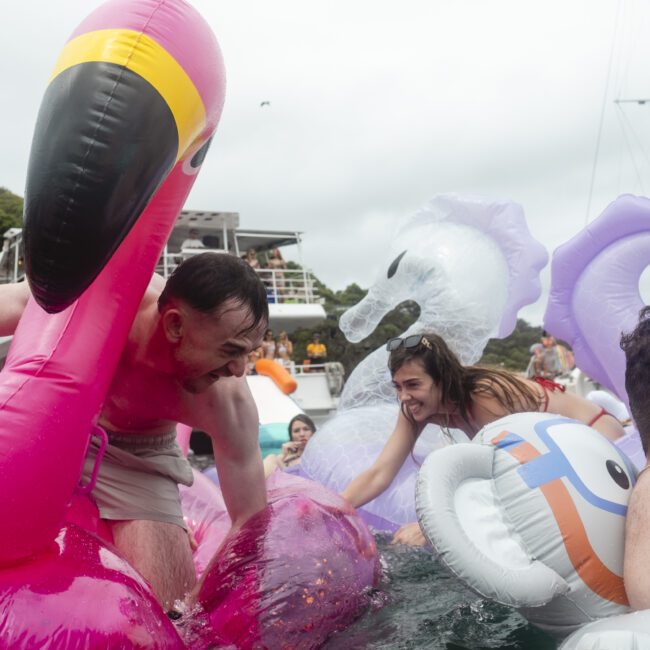 People are playfully climbing onto inflatable pool floats shaped like a flamingo and a unicorn in a body of water. A boat and trees are visible in the background. The atmosphere is lively and fun.