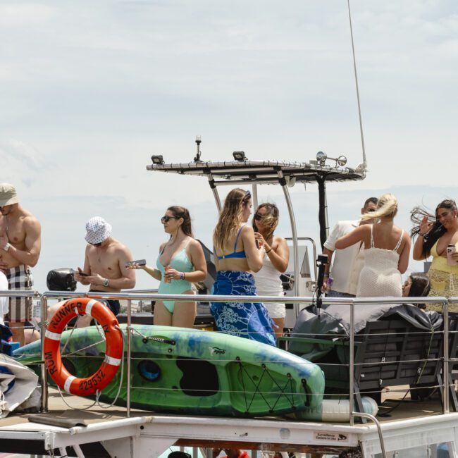 A group of people enjoying a sunny day on the deck of a boat. They are socializing and some are holding drinks. A kayak and life preserver are visible on the boat, with a calm water and sky in the background.