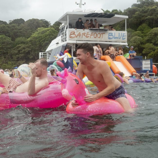 Groups of people enjoy a water party near boats. Some are riding inflatable toys, including a pink unicorn. The scene is lively, with lush green trees in the background and a sense of fun and relaxation.