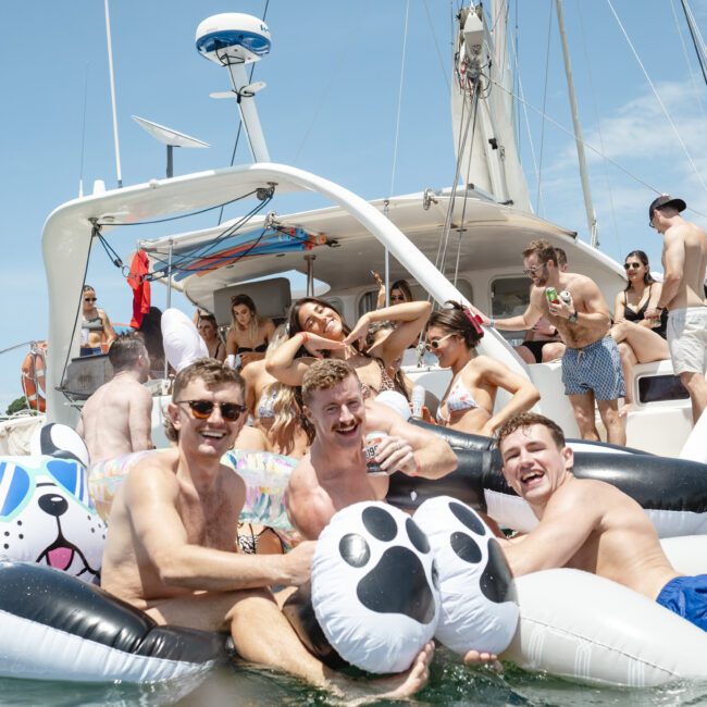 A group of people enjoying a sunny day on a catamaran and inflatable floaties shaped like animals. They're on a body of water, with some holding drinks and wearing swimsuits, capturing a festive and relaxed atmosphere.