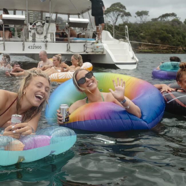 A group of people floating on colorful inflatables in the water, enjoying beverages and smiling. A boat is in the background with more people onboard. The setting is lively and festive, surrounded by natural scenery.