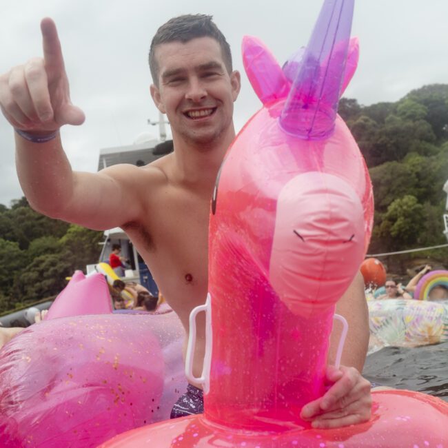 A man smiles as he sits on a pink inflatable unicorn in a body of water. He is pointing toward the sky, surrounded by trees in the background. Other colorful inflatables can be seen around him.