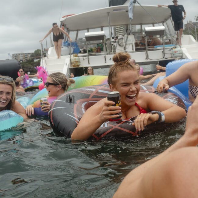 A group of people enjoying a party in the water near a yacht. They are on inflatable toys, laughing, and holding drinks. The sky is overcast, and a few people are on the deck of the yacht in the background.