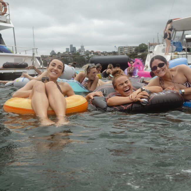 A group of people relax on inflatable rafts in a body of water, smiling and enjoying themselves. Boats are in the background under a cloudy sky. Some hold drinks, and the atmosphere is carefree and lively.