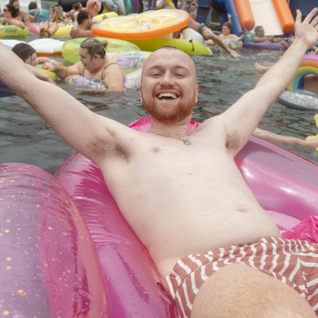 A man with a beard smiles and spreads his arms wide while lounging on a pink inflatable float in a pool. He is wearing striped swim trunks. In the background, people enjoy the water with various colorful inflatables.