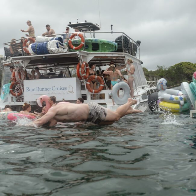 A group of people are enjoying a boat party on the water. Several individuals are swimming and floating on inflatables near the boat. The scene is lively and festive with a cloudy sky in the background.