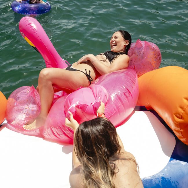 A woman in a bikini is laughing while reclining on a pink inflatable float. She's surrounded by others on a large floating platform in the water. The scene is sunny and playful, with blue-green water visible.