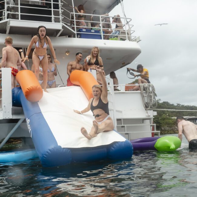 A group of people enjoying a boat party with a water slide. Two women in swimsuits are sliding into the water while others watch and cheer. A few people are swimming nearby, and a cloudy sky is overhead.