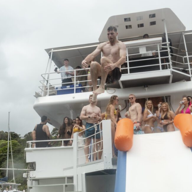 A shirtless man in mid-air slides off an inflatable slide on a boat. Onlookers cheer and take photos. The boat is anchored in a scenic area with greenery and a bird flying in the sky.