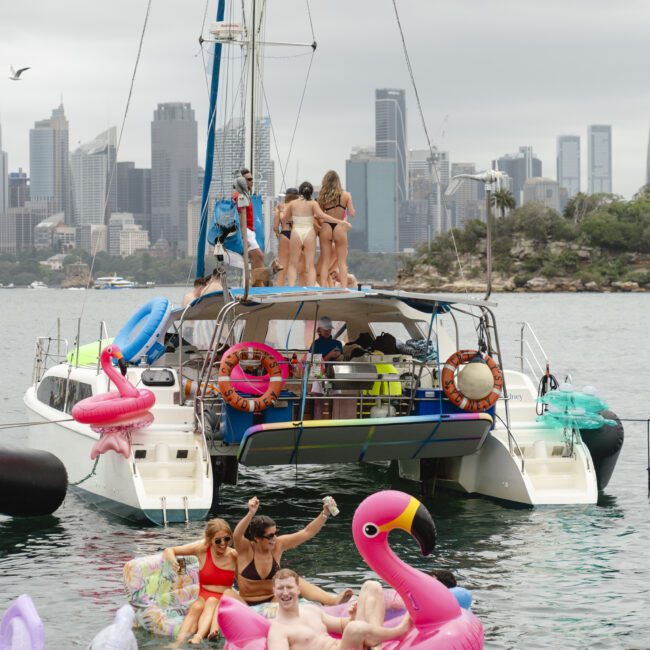 A group of people enjoying a day on a catamaran surrounded by inflatable pool toys in a harbor. The city skyline is visible in the background, with lush greenery on a nearby island. A bird flies overhead.