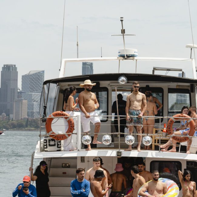 A group of people in swimwear enjoying a sunny day on a boat with city skyscrapers in the background. Some are standing, others are sitting or talking. Inflatable toys and life rings are visible on the deck.