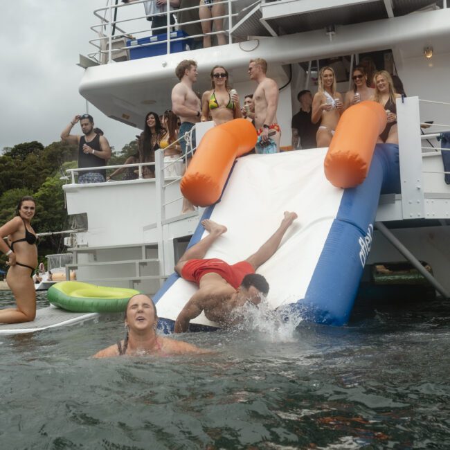 A group of people enjoying a boat party. Two individuals slide down an inflatable water slide into the sea, while others watch and socialize on the boat deck. The background has lush green trees and a sandy beach.