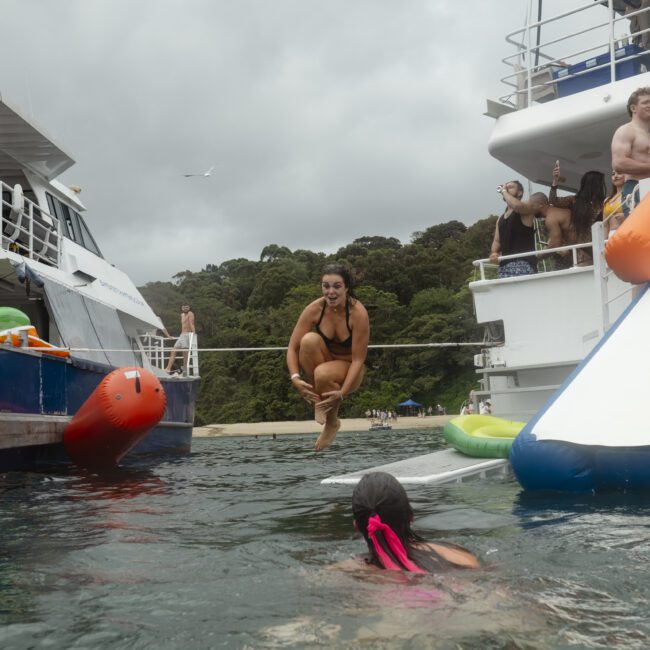 A woman is mid-air jumping from a boat into the water, surrounded by others on boats and a waterslide nearby. Some people in swimsuits watch her, and a seagull flies overhead against a cloudy sky, with a forested shoreline in the background.