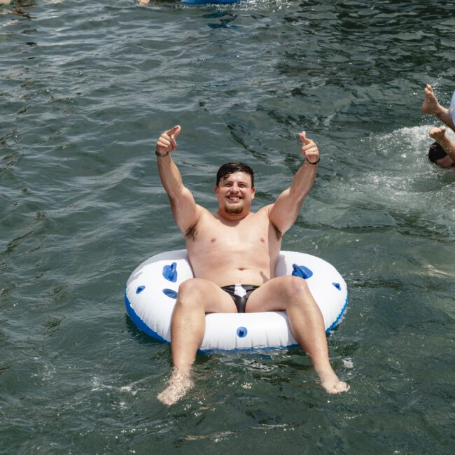 A man in a white inflatable ring gives two thumbs up while floating in a body of water. Other people are swimming and using inflatable rings in the background. The water is clear and the day appears sunny.