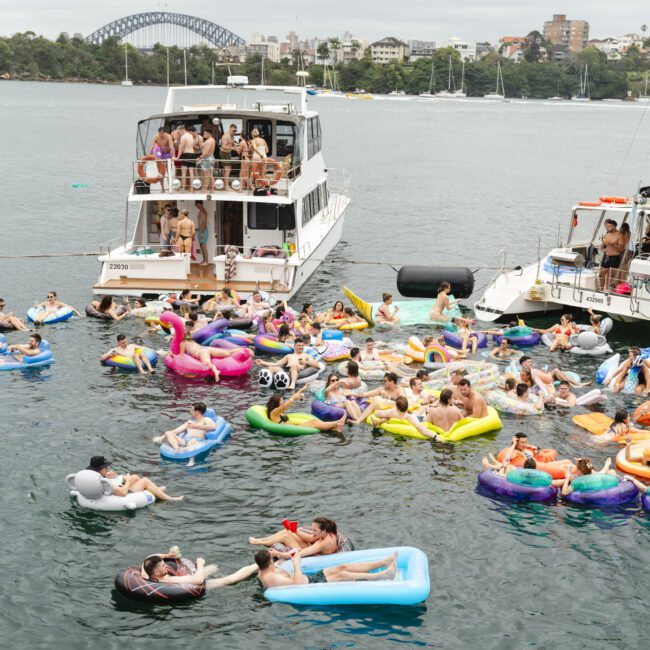 A lively scene with people enjoying a sunny day on colorful inflatable floats next to boats in the water. The background features a cityscape and a bridge under a cloudy sky.