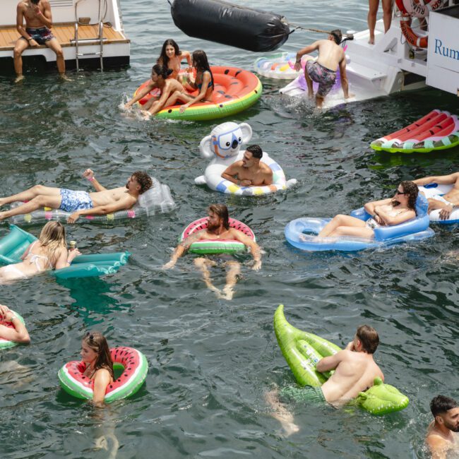 People relaxing on colorful pool floats in the water near a boat. Floats include a watermelon slice, a palm tree, and a swan. The scene is lively, with a backdrop of a boat and the calm water.