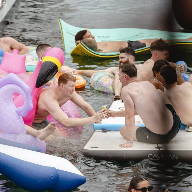 A group of people enjoying a pool party, lounging on inflatables, and sitting on a floating platform. They are holding drinks and socializing, surrounded by inflatable animals and pool toys. The atmosphere is lively and relaxed.