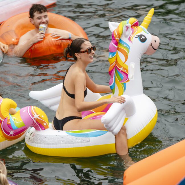 People enjoying a sunny day in a lake, sitting on colorful inflatable floats, including a unicorn. Some are holding drinks, and everyone is smiling and chatting. The water is calm and the atmosphere is festive.