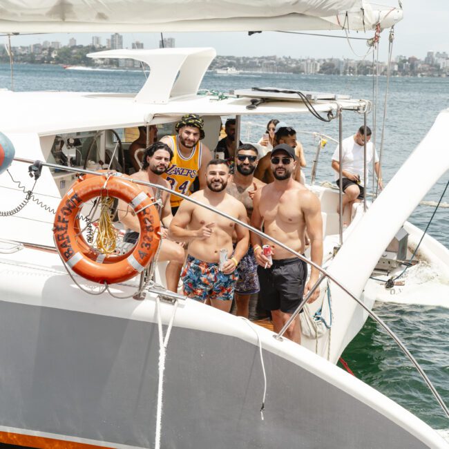 A group of seven smiling people stand on the deck of a sailboat, enjoying a sunny day on the water. They're casually dressed and showing thumbs up, with a city skyline visible in the background. A lifebuoy is attached to the boat's railing.