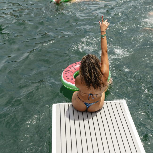 A woman in a swimsuit sits on the edge of a dock, holding up her hand. Several people float in the water on watermelon-themed inflatables, enjoying a sunny day. The water is calm and reflects the bright summer sky.