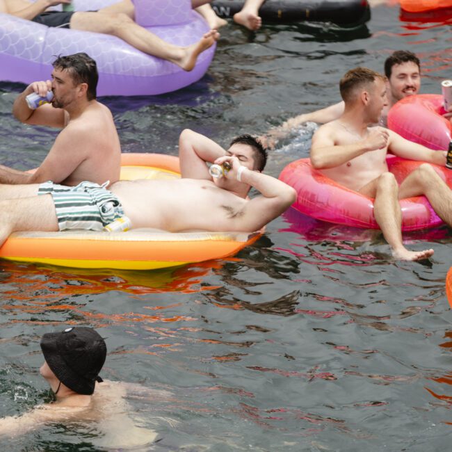 A group of men lounging on colorful inflatable floats in a body of water, holding drinks and relaxing. Some are sitting upright while others are lying down, enjoying a sunny day.