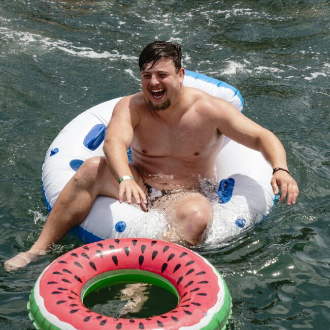 A man floating on a blue and white inflatable ring in a lake, smiling and splashing water. Another inflatable ring with a watermelon pattern is nearby. Other people and inflatables are visible in the background.