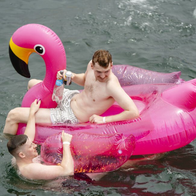 Two men are enjoying a day on the water with a giant pink inflatable flamingo. One man is sitting on the float sipping a drink, while the other playfully holds onto the flamingo in the water. The atmosphere is lively and fun.