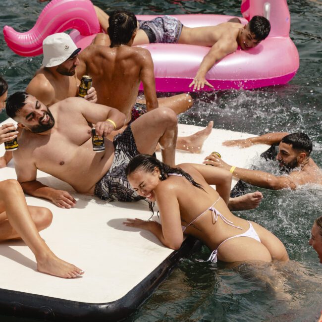 A group of people enjoying a sunny day on the water. Some are relaxing on a floating platform and a pink inflatable, while others swim. They are in swimwear, holding drinks, and smiling. The water is dark and rippling.