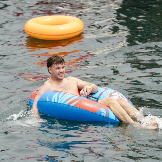 A person relaxes on a colorful inflatable ring in a body of water. An empty orange inflatable ring floats nearby. The person is smiling and enjoying the water.