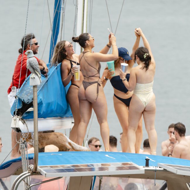 A group of people on a boat enjoying a sunny day. Four women in swimsuits stand together, holding drinks, with others seated around them. The scene is lively, with a crowd in the background and a calm body of water visible.