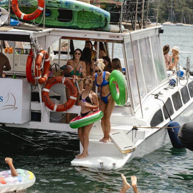 A group of people in swimwear enjoy a boat party on the water. Some are on the boat, holding inflatable rings, while others float nearby in the water. The background shows a city skyline and greenery.