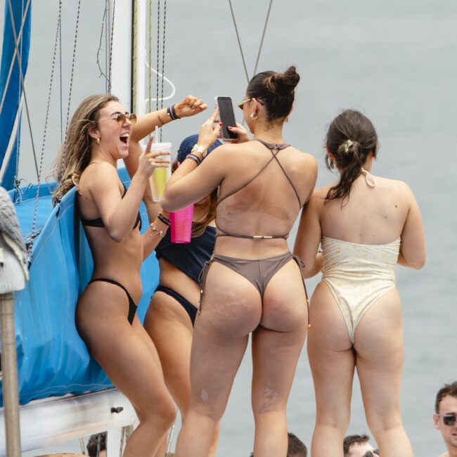 A group of women in swimsuits are gathered on a boat. One is taking a selfie with a phone while the others are smiling and holding drinks. The background shows a large body of water and part of the boat's structure.