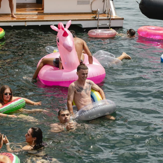 A group of people enjoy a summer day in the water, surrounded by colorful inflatable floats, including a pink unicorn. A boat is docked nearby, and everyone appears to be having a fun, relaxed time in the sun.