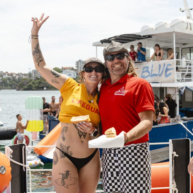 A smiling man and woman in swimwear stand on a dock near a boat named "Blue." The woman wears a yellow top, sunglasses, and has tattoos. The man wears a red shirt and checkered shorts. Both hold plates of food, and inflatables float on the water.