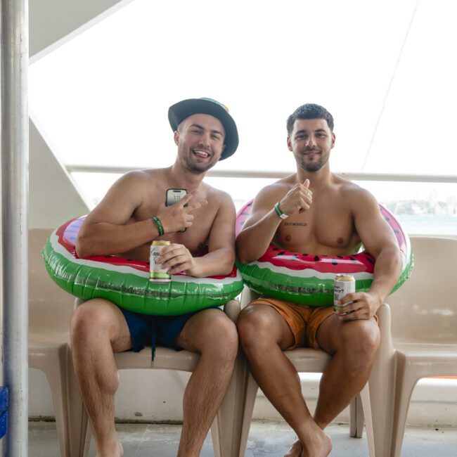 Two men sitting on a boat, wearing inflatable watermelon rings and swimsuits. They are smiling and holding drinks with thumbs up. Bright outdoor setting with a sea view in the background.