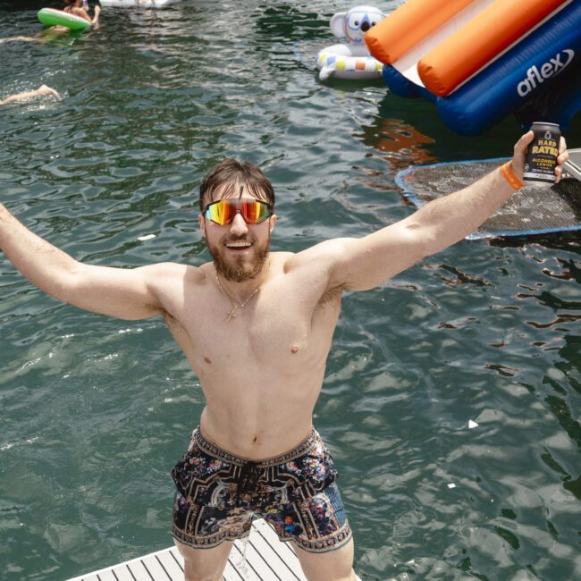 A man stands on a pier with arms raised, holding two beverage cans. He wears sunglasses and colorful shorts. Behind him, people enjoy the water, floating on inflatables. An inflatable slide is visible on the right. Fun, sunny atmosphere at a water event.