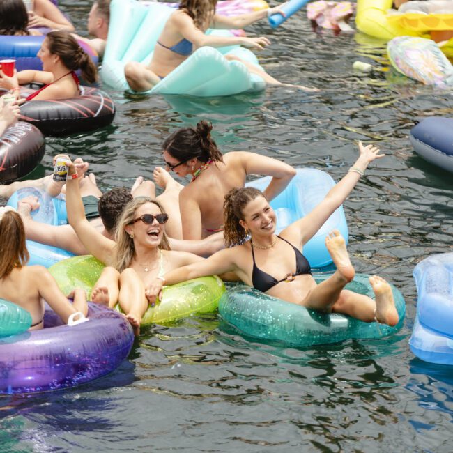 A group of people in swimsuits enjoy a sunny day on a river, lounging on colorful inflatable rings and floaties. They appear relaxed and cheerful, with some raising their arms in celebration, surrounded by clear water and other swimmers.