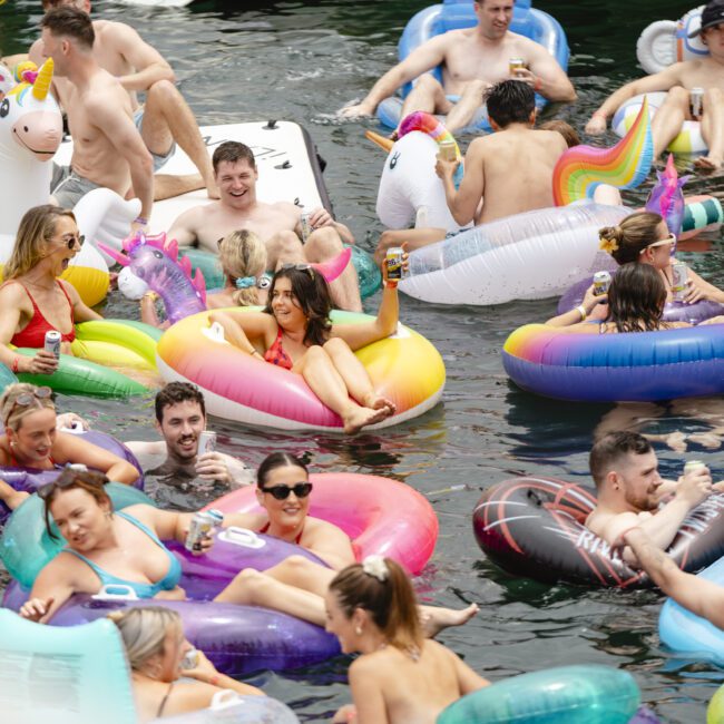 A large group of people enjoying a sunny day in the water, lounging on colorful inflatable rafts shaped like unicorns and other fun designs. They are smiling, holding drinks, and wearing swimwear, creating a lively, festive atmosphere.