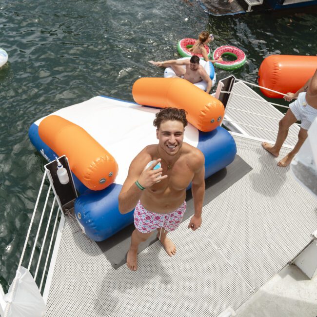 A man in swim trunks stands on a floating platform by the water, smiling and holding a drink. Colorful inflatables and people enjoying the water are in the background, creating a lively, summer atmosphere.