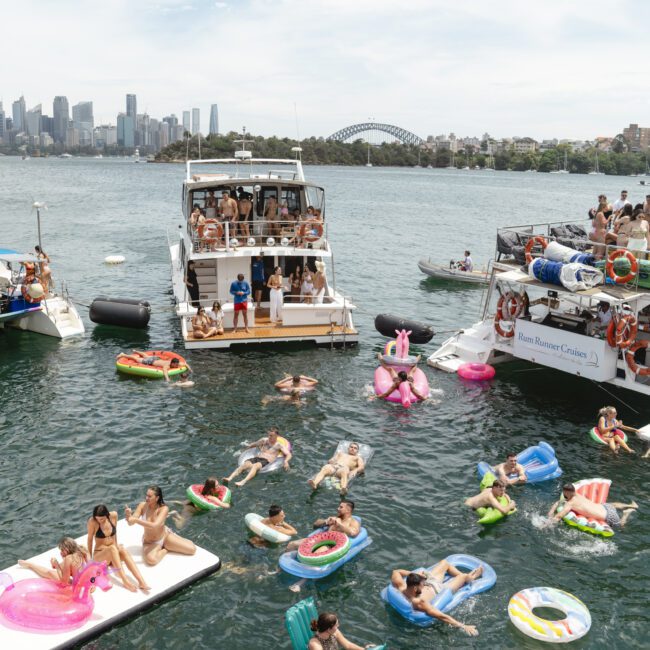 People are enjoying a sunny day on colorful inflatables in the water between three boats, with a city skyline and a bridge in the background. The scene is lively and festive, with clear skies above.