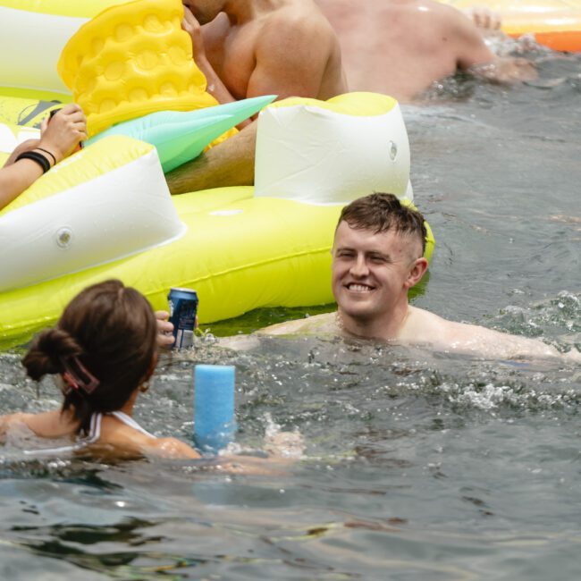 A group of people enjoying a swim in the water with inflatable floats. One person, holding a drink, is smiling while others relax nearby. Brightly colored inflatables add a cheerful atmosphere to the scene.