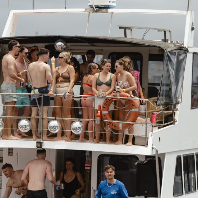 A group of people in swimsuits are gathered on the upper deck of a boat, socializing and holding drinks. Two lifebuoys are visible, and the sky is cloudy. A crew member stands on the lower deck.