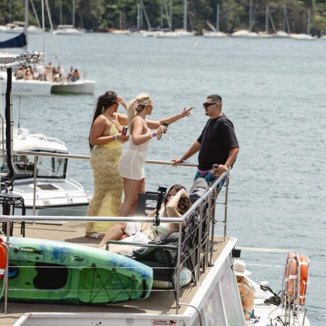 A group of people relaxing on a boat deck, with some standing and chatting, others lounging. A kayak is secured nearby. The background features a waterfront with trees and buildings under a clear sky.