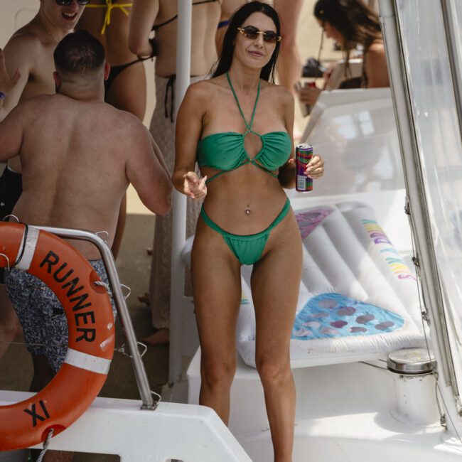 A woman in a green bikini stands on a boat, holding a drink. She is wearing sunglasses, and there are other people in swimwear socializing around her. A life preserver with "Riviera" on it hangs nearby.