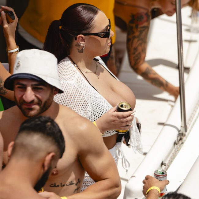 A group of people enjoying a party on a boat. Some are holding drinks and wearing casual summer attire. One person is wearing a white bucket hat and another is in a yellow sports jersey. Bright and sunny atmosphere.