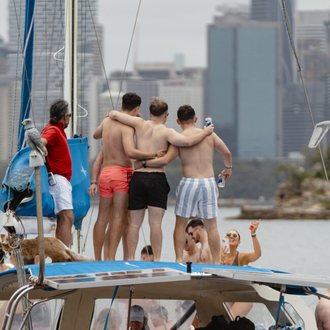 A group of people enjoying a day on a sailboat. Three men in swim trunks stand embracing on the deck, facing the city skyline. Others are seated or taking photos, with a dog resting nearby. The city buildings are visible in the background.