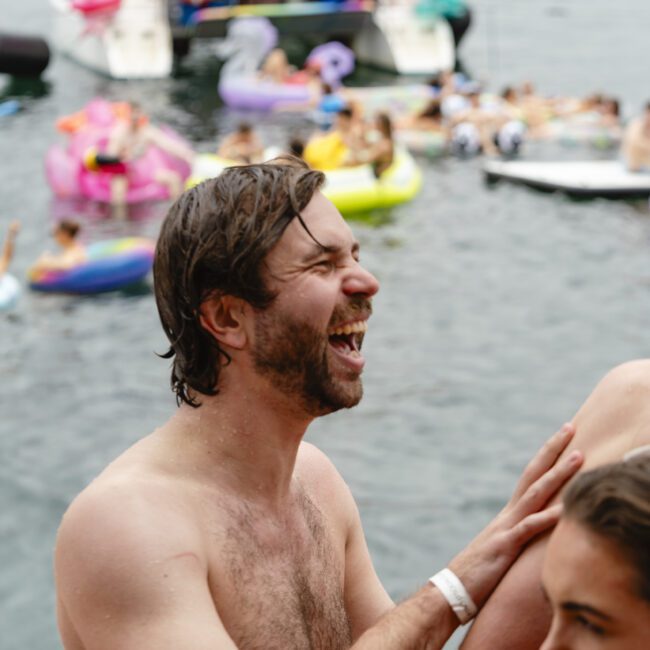 A man laughs while holding a drink on a boat. In the background, several people are relaxing on inflatables and boats in the water. The atmosphere is lively and festive.