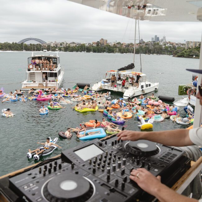 A DJ stands at a console on a boat overlooking a vibrant outdoor party. Dozens of people are floating on colorful inflatables in a body of water, surrounded by other boats and a city skyline in the background. The atmosphere is festive and lively.