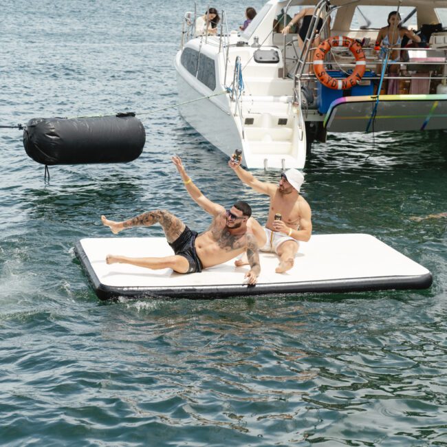 Two men relaxing on a floating platform in the ocean, smiling and waving. One holds a drink. There's a yacht in the background and another person swimming nearby. The sky is clear, and the mood is festive and cheerful.